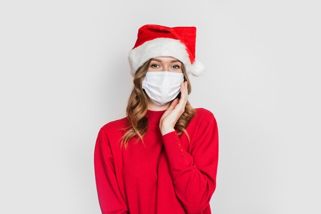 Het leuke Kaukasische studentenmeisje die een medisch masker en de rode sweater van de kerstmuts dragen houdt hand dichtbij gezicht dat op witte studioachtergrond wordt geïsoleerd