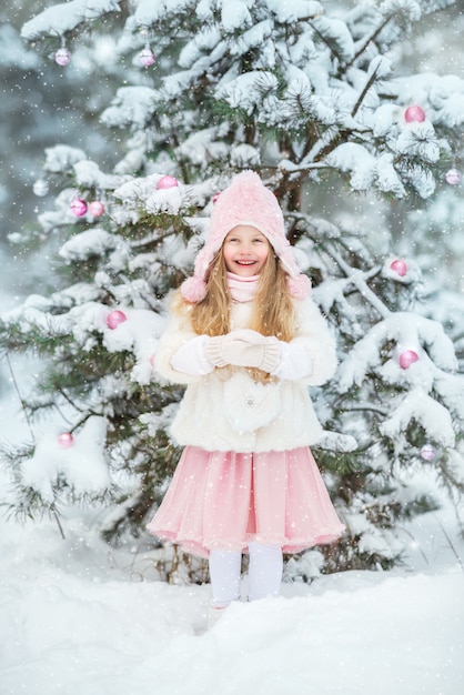 Het leuke meisje in een witte bontjas en een roze pluizige rok in het bos in de winter kleedt omhoog de Kerstboom. Roze Kerstspeelgoed.