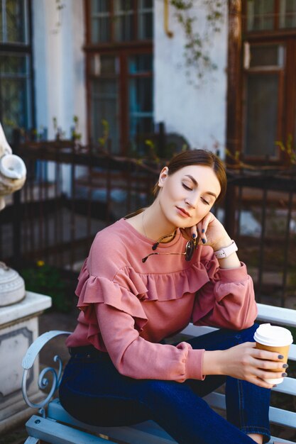 Het leuke nadenkende meisje in een roze sweater en jeans zit op een witte bank en houdt een kop van koffie