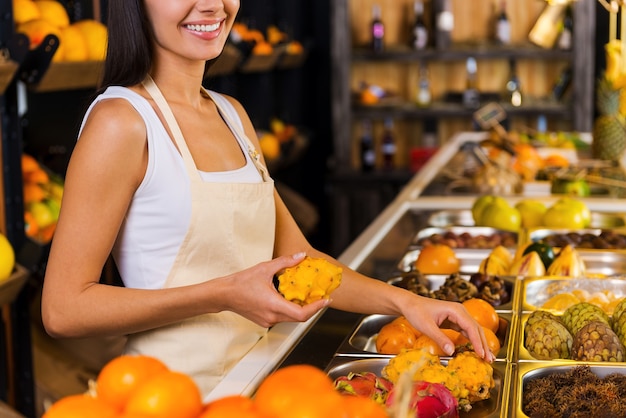 Het meest verse fruit voor u kiezen. Bijgesneden afbeelding van mooie jonge vrouw in schort die werkt in de supermarkt met verschillende soorten fruit op de achtergrond