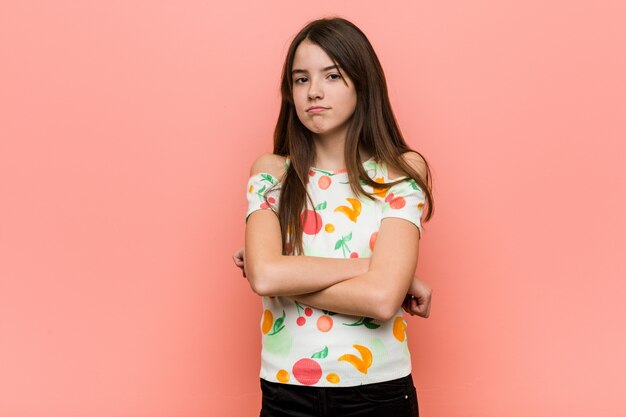 Het meisje dat de zomer draagt kleedt zich tegen een rood muur fronsende gezicht in ongenoegen, houdt wapens gevouwen.