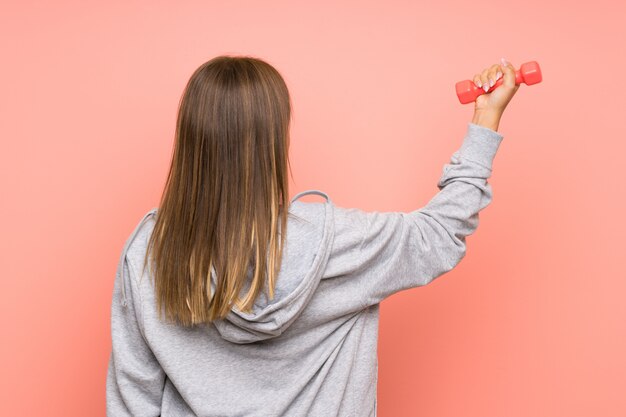 Het meisje dat van de tienersport gewichtheffen over geïsoleerde roze muur in achterpositie maakt