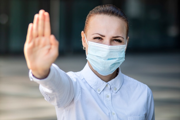Het meisje, de jonge vrouw, de onderneemster in beschermend steriel medisch masker op gezicht, tonen palm, hand, ophouden geen teken. Luchtverontreiniging, virus, pandemisch coronavirus concept. Covid-19