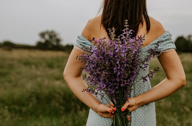 Het meisje houdt in haar handen een groot boeket lila en violette bloemen achter haar rug