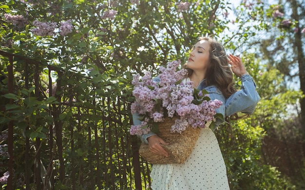 Het meisje houdt in haar handen een rieten mand met bloemen. Mand met seringen. Meisje en bloemen. Loop met een mand met seringen in de handen. Floristiek. Lente lila bloesem