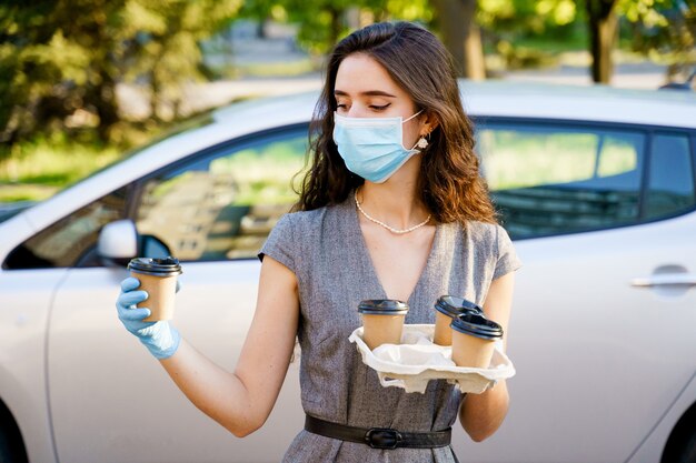 Het meisje in medisch masker en handschoenen houdt dienblad met wegwerpkoffiekopjes