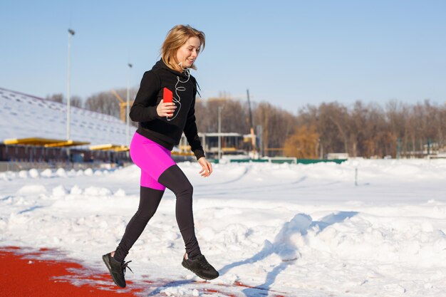Het meisje in sportkleding loopt op het rode spoor voor het lopen op een met sneeuw bedekt stadion Pasvorm en sportlevensstijl. Ren en luister muziek. Sport levensstijl