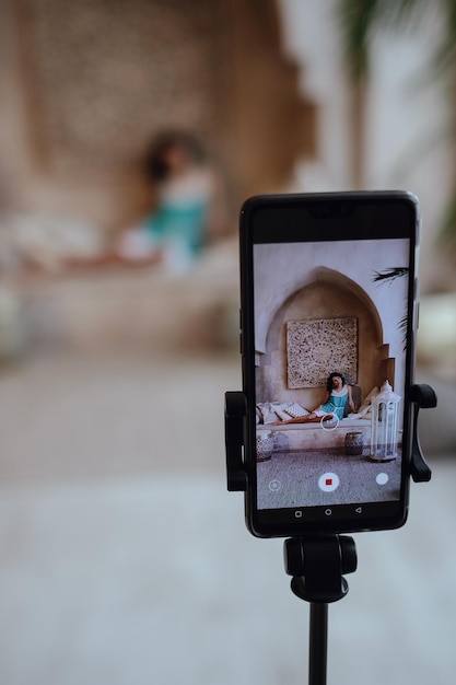 Het meisje is gefotografeerd op een smartphone die op een statief staat in een harem in Marokkaanse stijl. Charmant model poseren in een rode jurk tijdens haar vakantie naar Marokko in haar riad in Marrakech