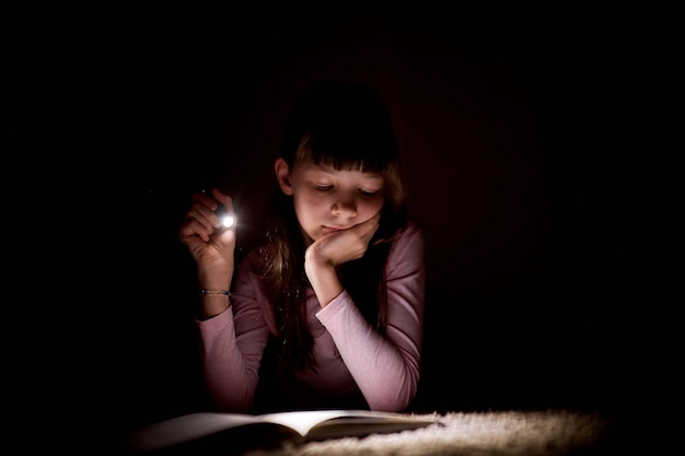 Het meisje leest een boek met een flitslicht in een donkere ruimte bij nacht.