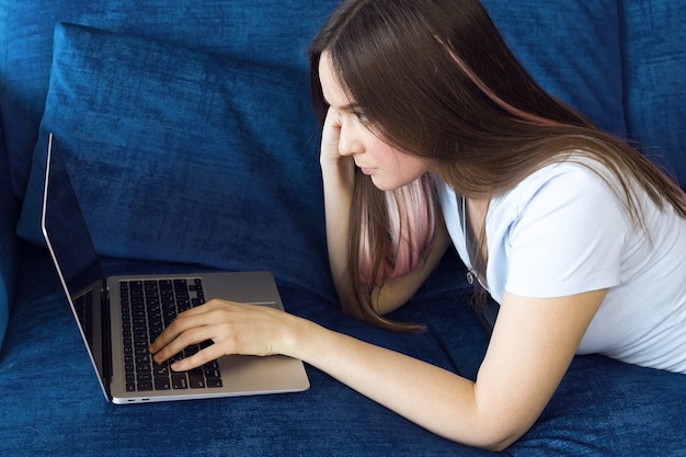 Het meisje ligt op de bank en werkt achter de laptop