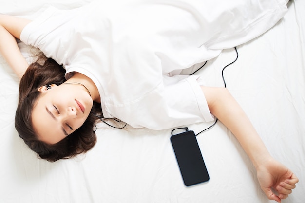 Het meisje ligt op het bed met een smartphone, luistert naar de radio met zijn ogen dicht.