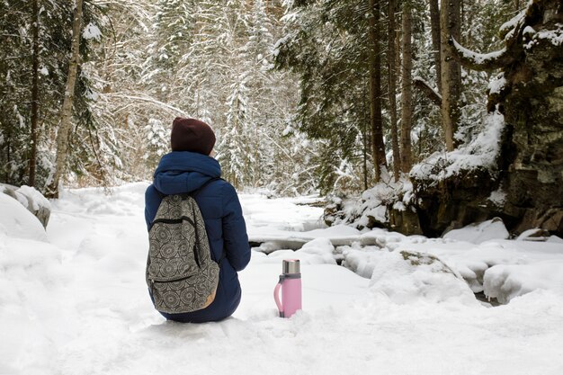 Het meisje met rugzak en thermosfles zit in een sneeuw naaldbos.
