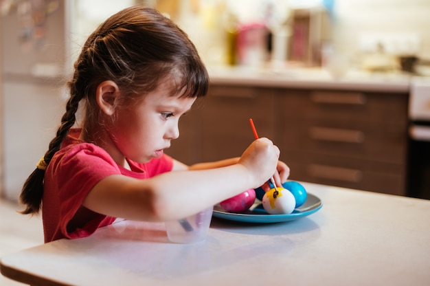 Het meisje schildert paaseieren op blauwe schotelzitting bij de lijst in keuken