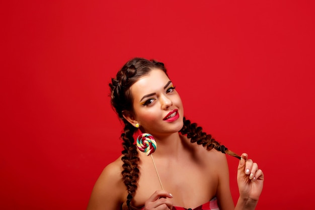 Het meisje van de schoonheidsmannequin die kleurrijke lolly eten. Verrast grappige jonge vrouw met vlechten kapsel, nagels en beige make-up, geïsoleerd op een rode muur