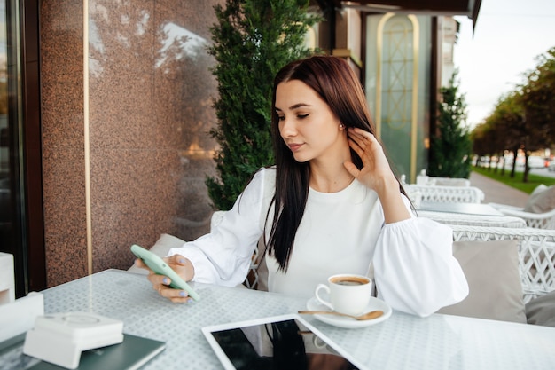 Het meisje zit in een café met behulp van technologie. het meisje kijkt naar een teleon in een café aan een tafel.