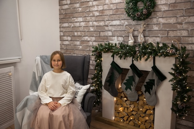 Het meisje zit in een stoel bij de open haard in de woonkamer, kerstinterieur