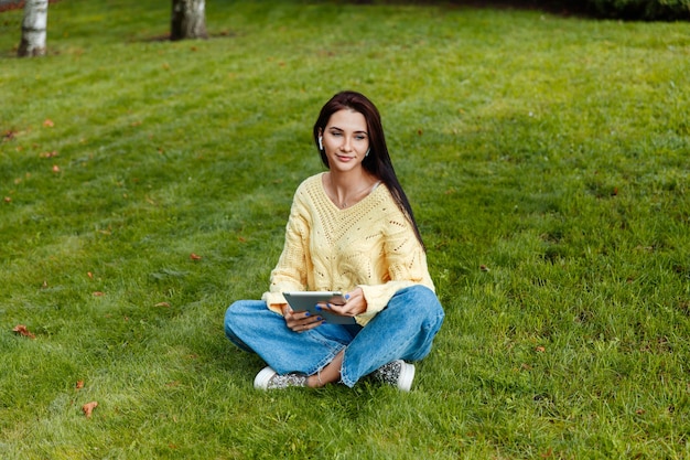 Het meisje zit in het park en is bezig met de iPad. jonge brunette zit op het gras en kijkt naar de tablet. meisje in het herfstpark.