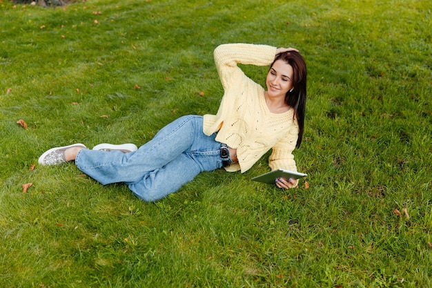 Het meisje zit in het park en is bezig met de ipad. jonge brunette zit op het gras en kijkt naar de tablet. meisje in het herfstpark.