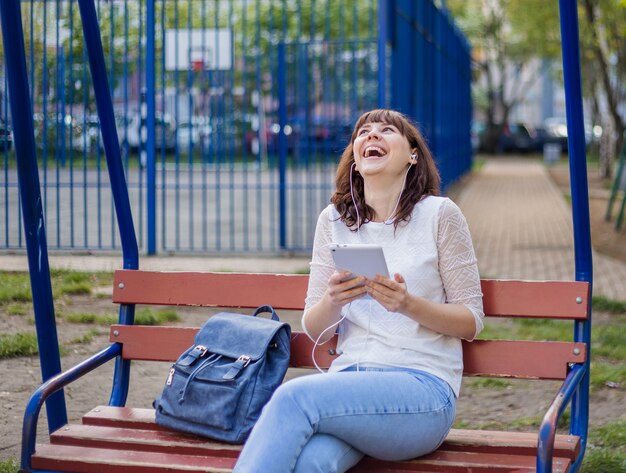 Het meisje zit op een bankje met een tablet en lachen, opzoeken. brunette meisje in een wit jasje en jeans op straat