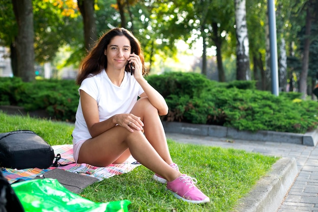 Het meisje zit op het gras in het park en communiceert aan de telefoon