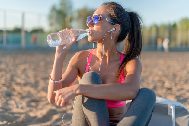 Het mooie de vrouwen drinkwater van de geschiktheidsatleet na werkt het uitoefenen op de zomer van de zonsondergangavond in strand openluchtportret uit.