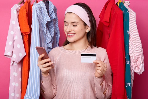 Het mooie donkerharige jonge wijfje met vrolijke uitdrukking, houdt slimme telefoon en creditcard. Gelukkig meisje voert online betaling uit. Vrouw kiest outfits in online shop. Betaling en winkelen concept.