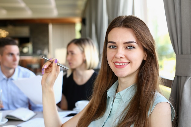 Het mooie glimlachende vrolijke meisje op het werk kijkt in camera met collega'sgroep. Administratieve arbeider bij werkruimte, baanaanbieding, cliëntbezoek, het concept van de beroepstrein