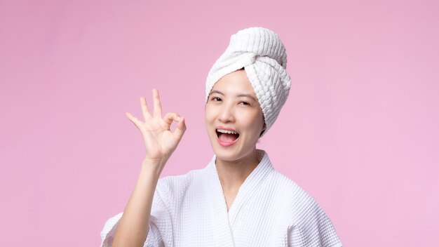 Het mooie jonge aziatische gezicht van de vrouwen gelukkige glimlach met hand of vingergebaartekens in badjas en douchehanddoek die lege ruimte tonen die op roze achtergrond wordt geïsoleerd