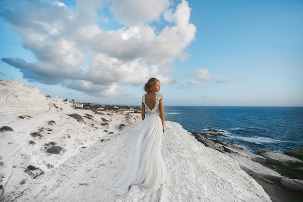 Het mooie jonge blonde modelmeisje, in witte kleding, gaat achteruit op de witte rots en kijkt naar de zee