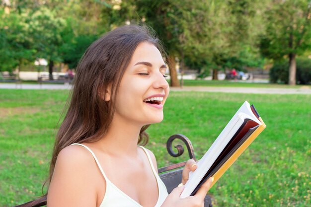 Het mooie jonge brownhairmeisje ruikt een verse gedrukte boekzitting op de bank in het park