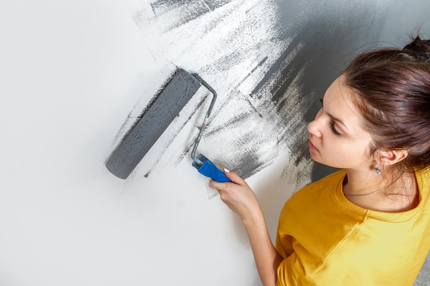 Het mooie jonge meisje schildert een muur in een geel jasje met een rol, grijze verf. Het concept van reparatie, verandering, ontwerp, interieur.