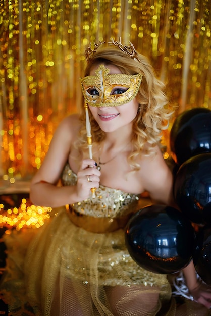 Het mooie jonge meisjes modelblonde glimlachen behandelt haar gezicht met een Carnaval-masker in een elegante gouden kleding met een hoepel hoornen achtergrond van lotuslinten met slingers die op de vloer zitten.