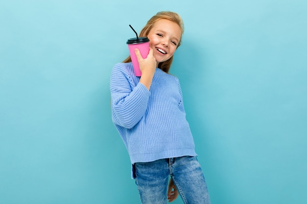 Het mooie Kaukasische tienermeisje met bruin haar in blauwe hoody drinkt koffie met roze die kop op blauw wordt geïsoleerd