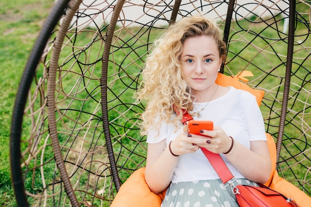 Het mooie krullende wijfje houdt slimme telefoon, typt tekstbericht, heeft goede rust in hangende stoel openlucht