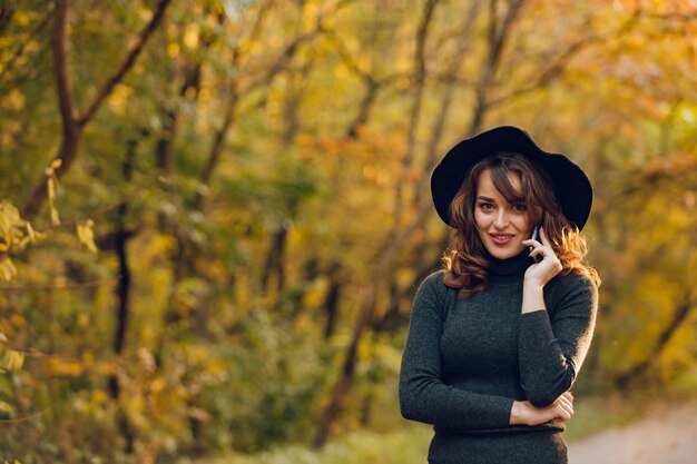 Het mooie meisje in een zwarte hoed houdt een telefoon in haar het park in de herfst in.