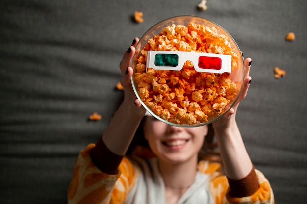 Het mooie meisje in pyjama eet popcorn, ligt op een grijze achtergrond in 3d glazen en bekijkt een film