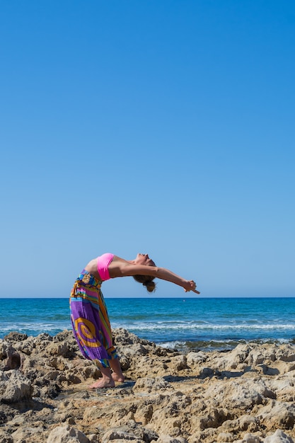 Het mooie meisje met een sportief figuur beoefent yoga buitenshuis in de buurt van de zee. Zomer.