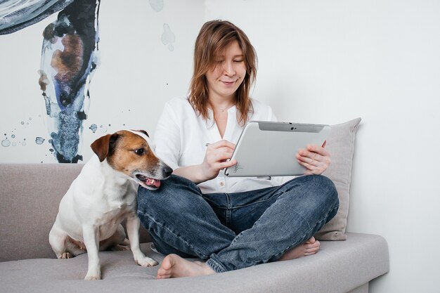 Het mooie meisje zit thuis op de bank met een tablet in haar handen, naast een Jack Russell-hond