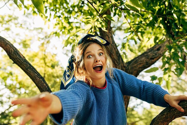 Het onderste weergavebeeld van een jonge blonde vrouw heeft plezier op de boom op de natuurachtergrond in het park Gelukkige vrouwelijke zitting gon de tak Mensen reizen levensstijl