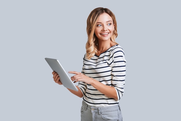 Het onderzoeken van haar nieuwe digitale tablet. Aantrekkelijke jonge vrouw die lacht en haar digitale tablet gebruikt terwijl ze tegen een grijze achtergrond staat