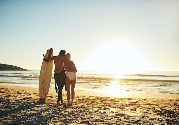 Het paar dat samen surft, blijft bij elkaar Achteraanzicht van een onherkenbaar jong stel dat staat en elkaar vasthoudt op het strand bij zonsondergang