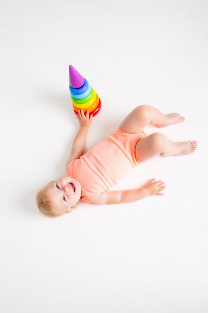 Het peutermeisje in oranje bodysuit ligt naast een veelkleurige piramide op een witte muur, mening van hierboven