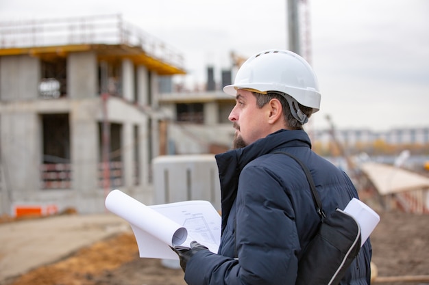 Het portret van architect aan het werk met helm op een bouwwerf, leest het plan, document projecten