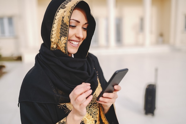 Het portret van charmante glimlachende moslimvrouw kleedde zich in traditionele slijtage gebruikend slimme telefoon terwijl buiten status.