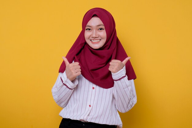 Het portret van de aziatische jonge vrouw die hijab draagt, geeft twee duimen en kijkt gelukkig uitdrukking