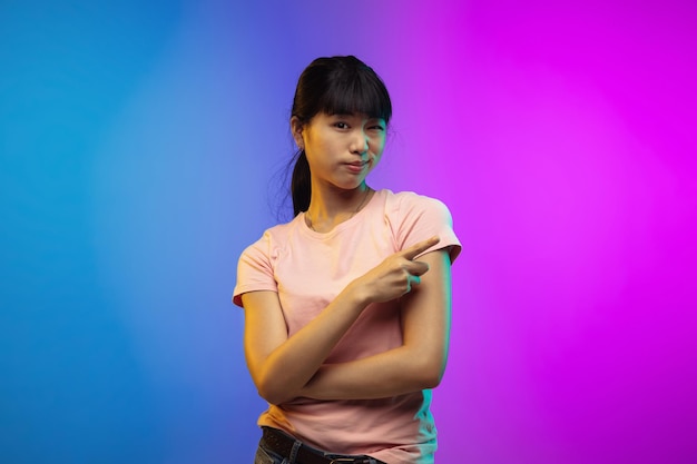 Het portret van de Aziatische jonge vrouw op de achtergrond van de gradiëntstudio in neon. Mooi vrouwelijk model in casual stijl. Concept van menselijke emoties, gezichtsuitdrukking, jeugd, verkoop, advertentie. Folder