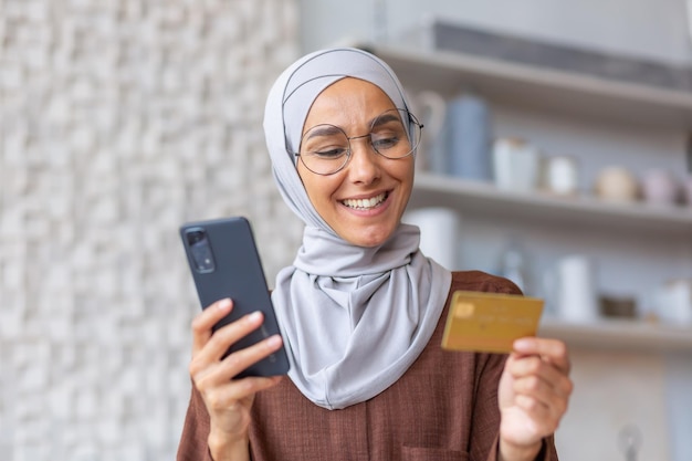 Het portret van de close-upfoto van jonge glimlachende Arabische vrouw in hijab thuis in keukenholding telefoon en