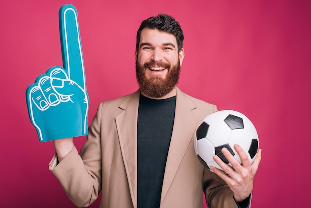 Foto het portret van de gebaarde mens houdt een voetbal of een voetbalbal en een nummer één handschoen van het ventilatorschuim op roze muur