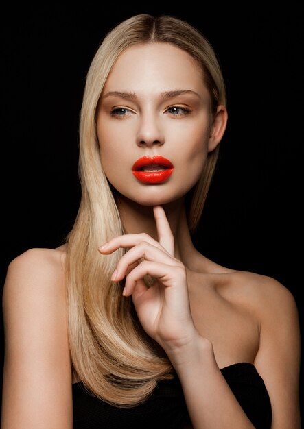 Het portret van de schoonheidsmannequin met glanzend blondekapsel met rode lippen op zwarte achtergrondholdingsvinger naast haar kin