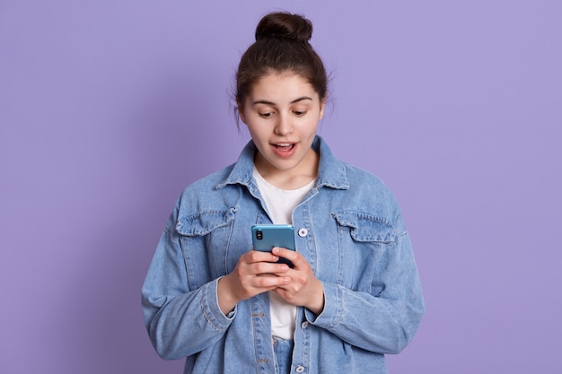 Het portret van de verbaasde vrouw met moderne slimme telefoon in handen, kleedt modieuze uitrusting, bekijkend het scherm van mobiele telefoon met verbaasde uitdrukking, geïsoleerd over lilac muur.